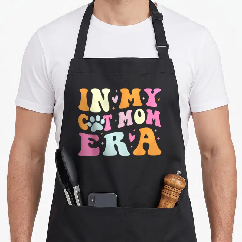 Funny era cat apron - Stacked retro text In My Cat Mom Era with paw print and pastel colors — shown on a black cotton canvas apron against a white background