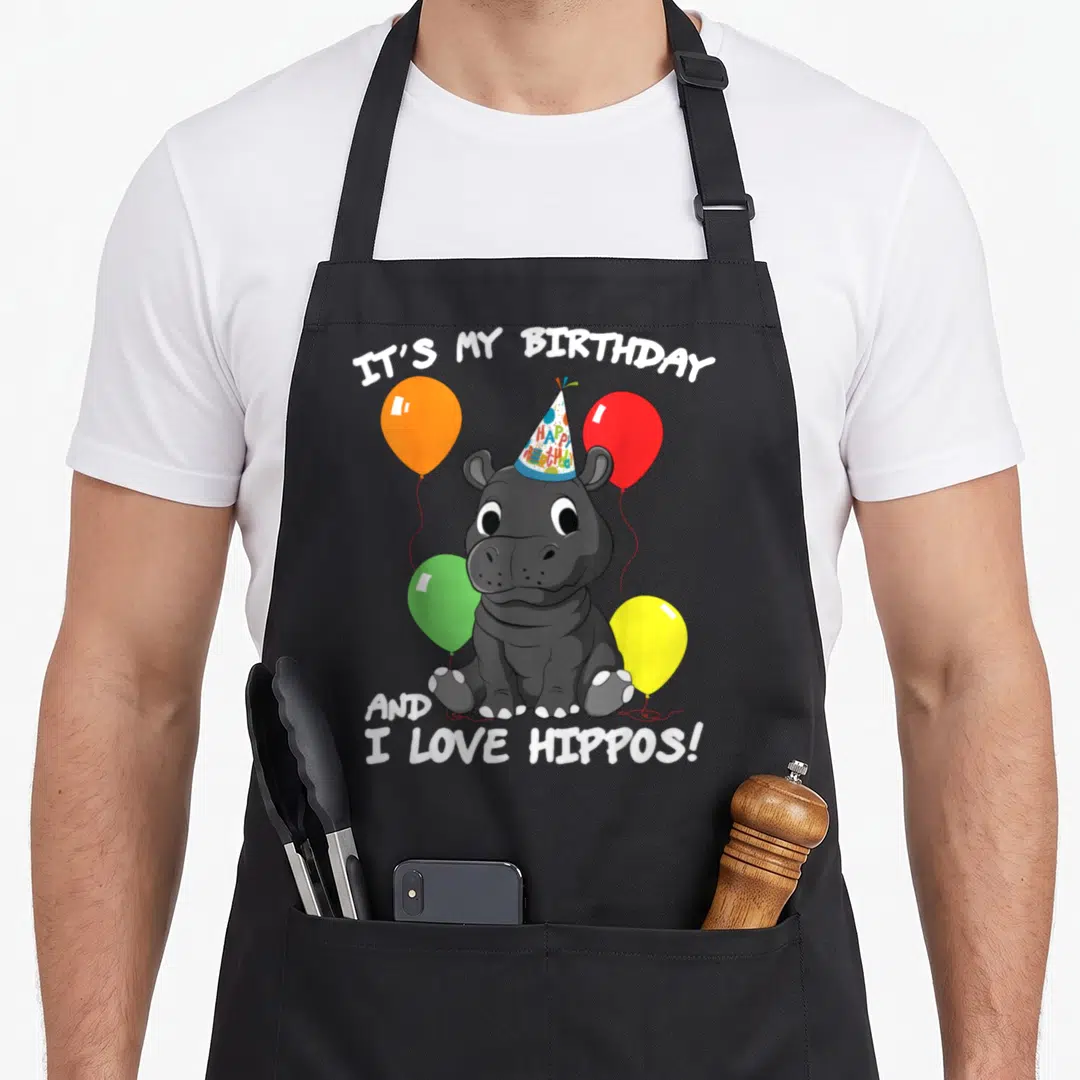 Hippo birthday apron - Grey cartoon hippo in party hat with four orange red green and yellow balloons and white — shown on a black cotton canvas apron against a white background