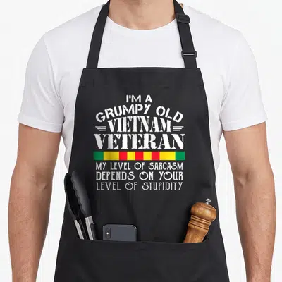 Underestimate vietnam veterans apron - Distressed white text reading I'm a grumpy old Vietnam veteran above a green — shown on a black cotton canvas apron against a white background