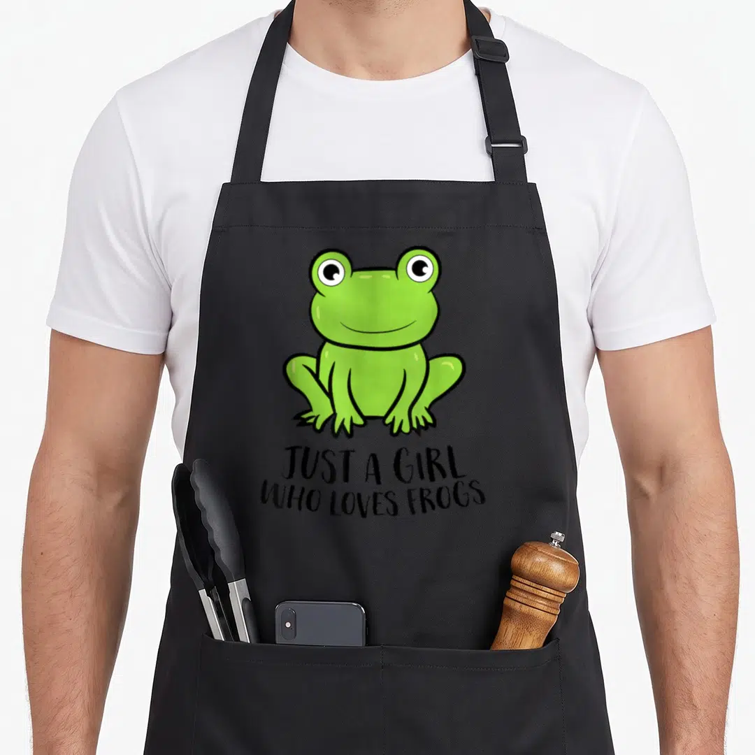 Vintage frog aprons - Cartoon green frog illustration above black text reading JUST A GIRL WHO LOVES FROGS — shown on a black cotton canvas apron against a white background