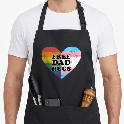 Free rainbow apron - Distressed heart split between rainbow and trans flags with stacked black text reading Free Dad — shown on a black cotton canvas apron against a white background