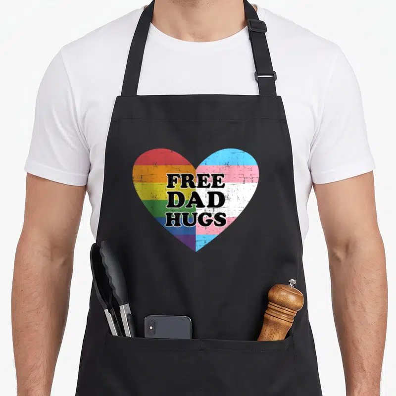 Free rainbow apron - Distressed heart split between rainbow and trans flags with stacked black text reading Free Dad — shown on a black cotton canvas apron against a white background