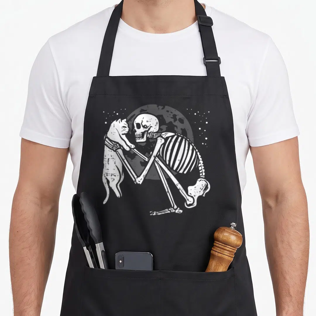 Kitty goth apron - White skeleton sitting and hugging a cat in front of a grey moon with stars — shown on a black cotton canvas apron against a white background