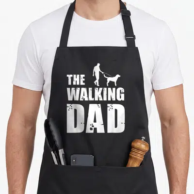 Walking top dog aprons - White silhouette of man walking dog above stacked text The Walking Dad — shown on a black cotton canvas apron against a white background