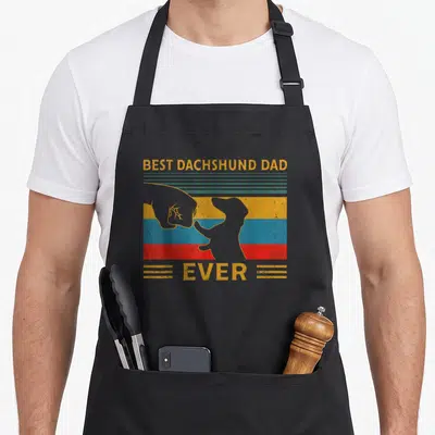 Weiner dog dad apron - Retro text Best Dachshund Dad Ever with hand and paw silhouettes over yellow blue red stripes — shown on a black cotton canvas apron against a white background