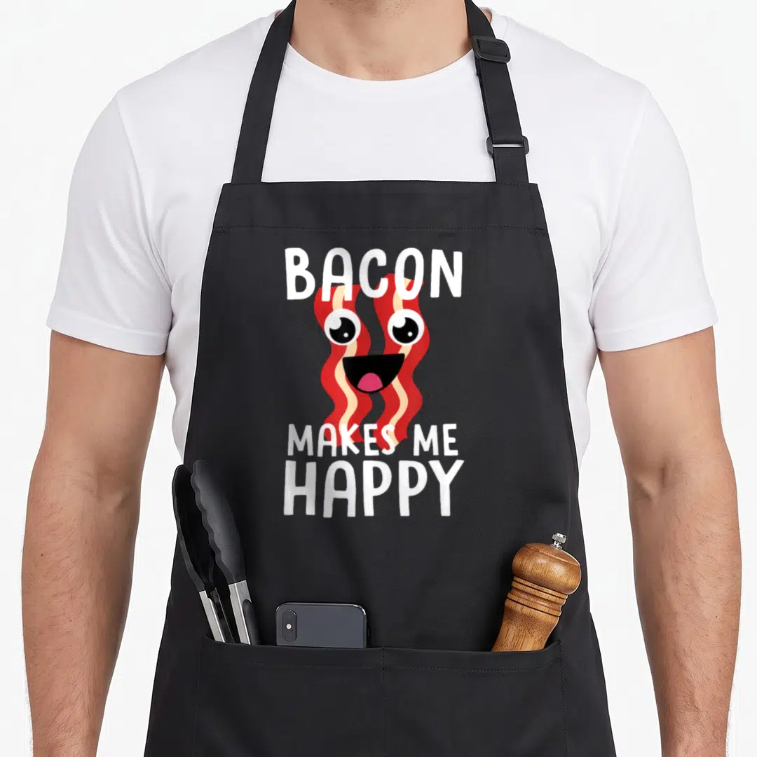 Cute bacon apron - Cartoon bacon strip with eyes and smile between white text BACON MAKES ME HAPPY — shown on a black cotton canvas apron against a white background