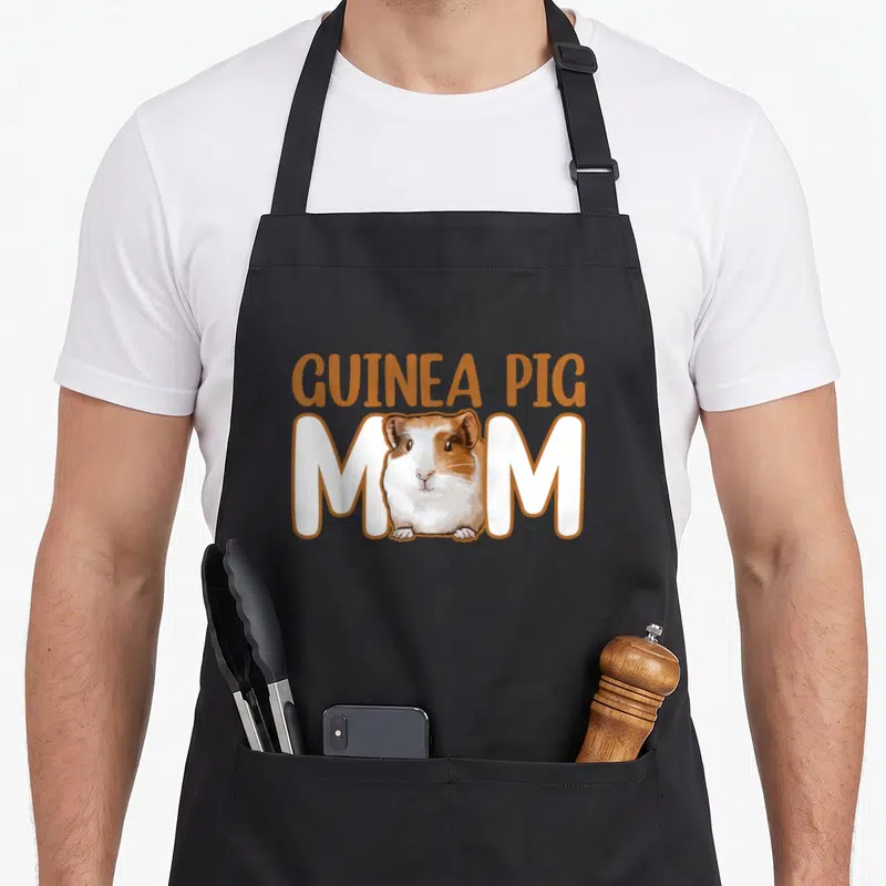 Cavy guinea mom apron - Cartoon tricolor guinea pig sits between white M letters under arched orange GUINEA PIG text — shown on a black cotton canvas apron against a white background