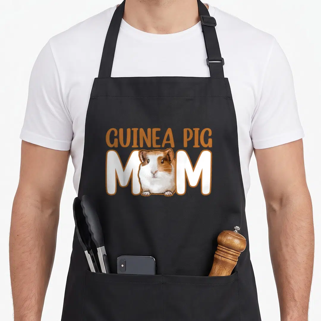 Cavy guinea mom apron - Cartoon tricolor guinea pig sits between white M letters under arched orange GUINEA PIG text — shown on a black cotton canvas apron against a white background