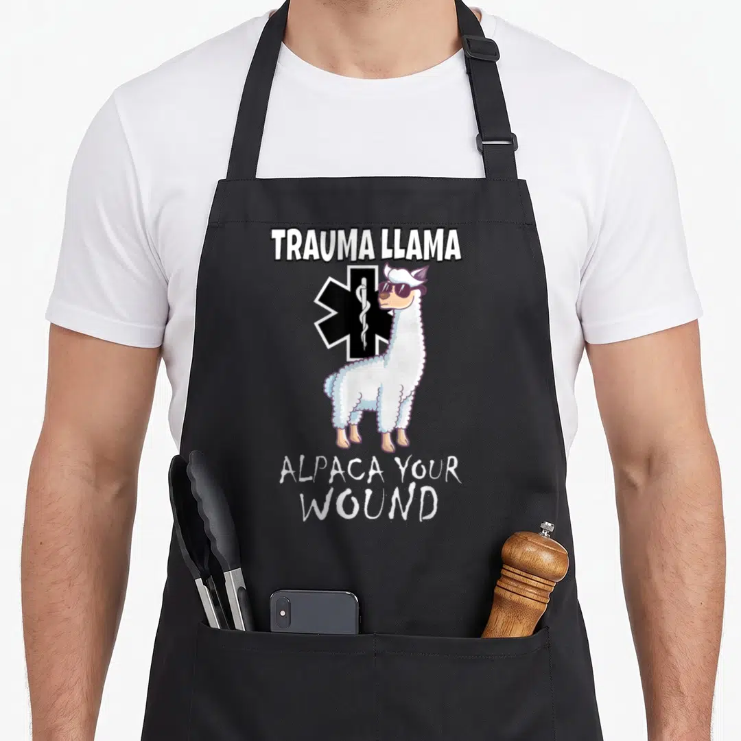 Trauma alpaca apron - Cartoon llama with sunglasses in front of Star of Life and text TRAUMA LLAMA ALPACA YOUR WOUND — shown on a black cotton canvas apron against a white background