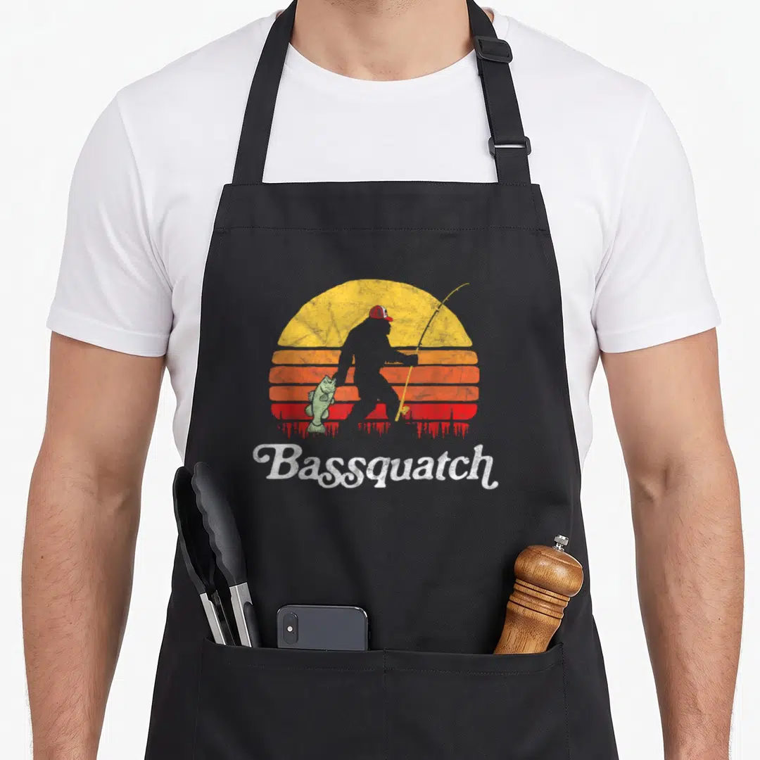 Bassquatch outdoor apron - Bigfoot silhouette holding fish and rod before striped retro sun with Bassquatch text — shown on a black cotton canvas apron against a white background