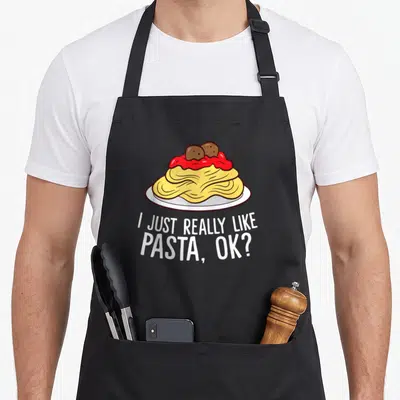 Funny pasta apron - Cartoon pasta plate with red sauce and meatballs above white text I JUST REALLY LIKE PASTA OK — shown on a black cotton canvas apron against a white background