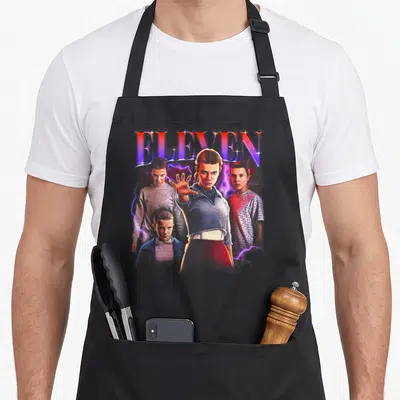 Heartthrob novelty apron - ELEVEN text above girl with outstretched hand, three boys, and purple lightning — shown on a black cotton canvas apron against a white background
