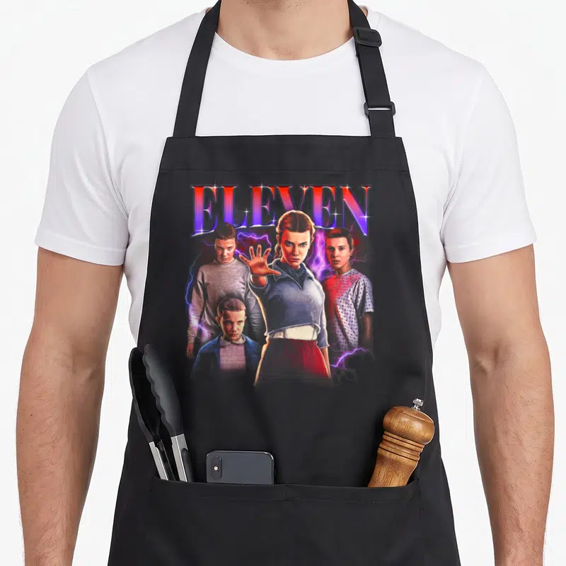 Heartthrob novelty apron - ELEVEN text above girl with outstretched hand, three boys, and purple lightning — shown on a black cotton canvas apron against a white background