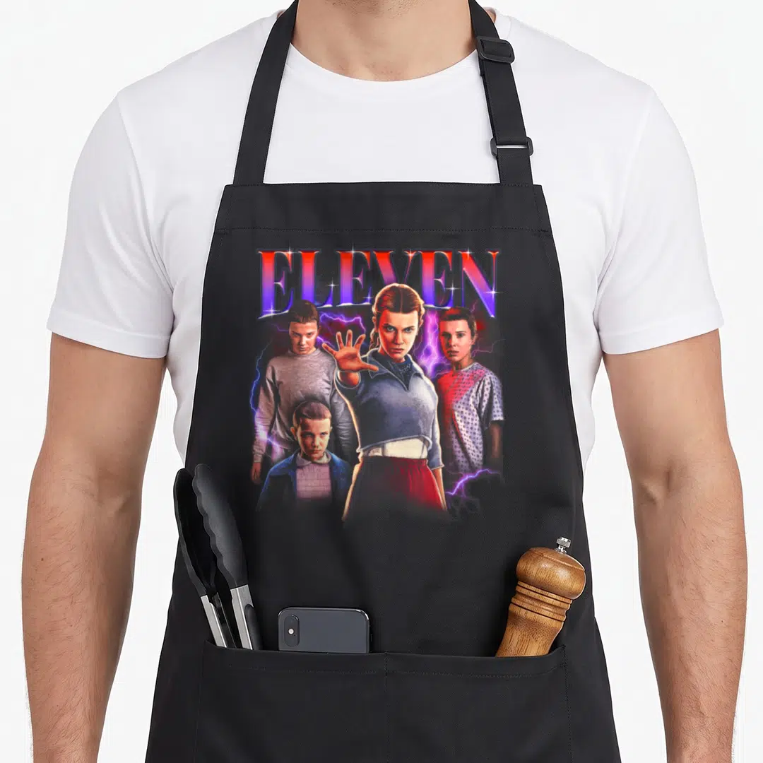 Heartthrob novelty apron - ELEVEN text above girl with outstretched hand, three boys, and purple lightning — shown on a black cotton canvas apron against a white background