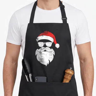 Hipster santa aprons - Stylized red hat white sunglasses and beard forming a minimalist face — shown on a black cotton canvas apron against a white background