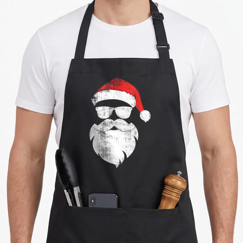 Hipster santa aprons - Stylized red hat white sunglasses and beard forming a minimalist face — shown on a black cotton canvas apron against a white background