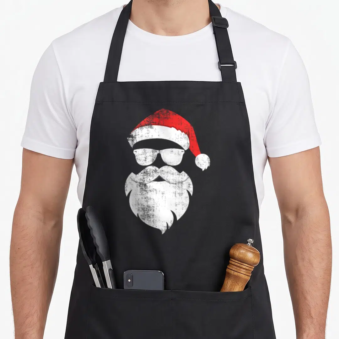 Hipster santa aprons - Stylized red hat white sunglasses and beard forming a minimalist face — shown on a black cotton canvas apron against a white background