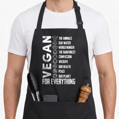 Vegan bee aprons - Vertical VEGAN text with nine icons and phrases about animals, hunger, and planet health — shown on a black cotton canvas apron against a white background