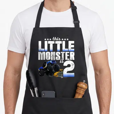 Monster 2nd birthday truck apron - Blue monster truck crushing yellow car next to number 2 and stacked text this LITTLE — shown on a black cotton canvas apron against a white background