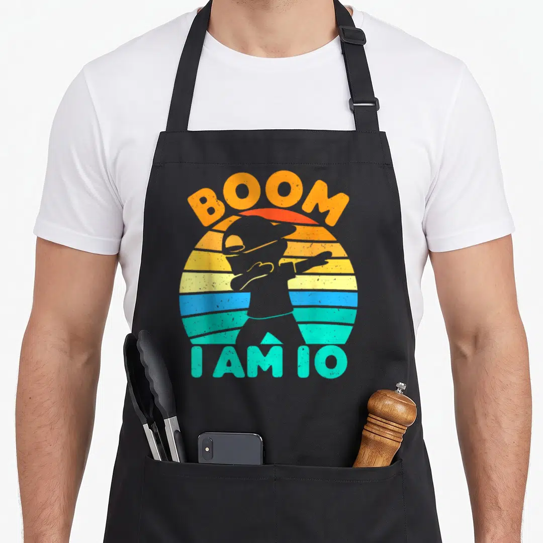 Boom birthday apron - Retro boy silhouette dabbing in front of striped sun with BOOM I AM 10 text — shown on a black cotton canvas apron against a white background