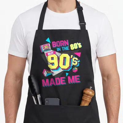 But 80s apron - Born in the 80s But 90s Made Me text with cassette tape, triangles, and cube in pink yellow blue white — shown on a black cotton canvas apron against a white background
