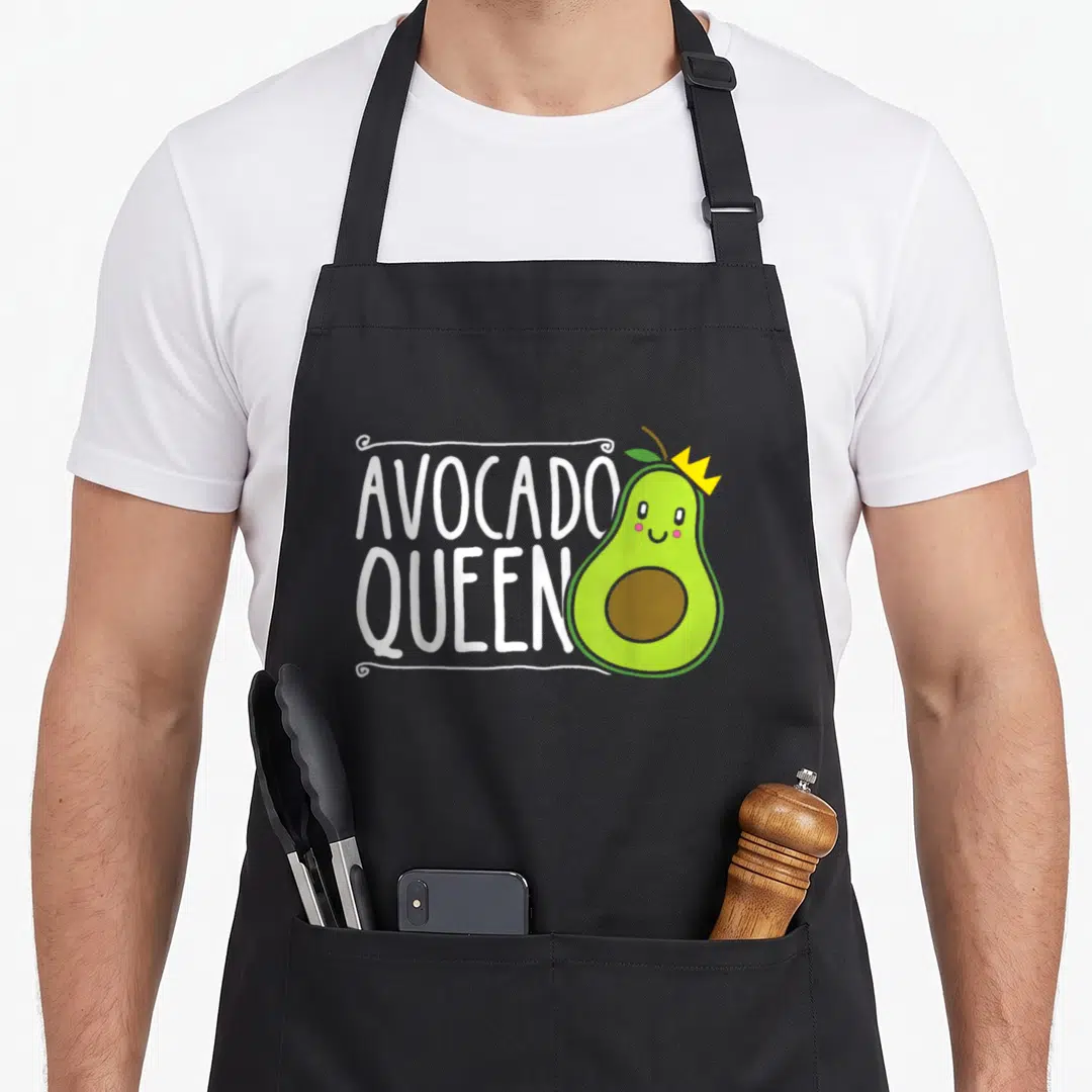 Cute queen avocado apron - Cartoon green avocado with yellow crown next to stacked white AVOCADO QUEEN text inside — shown on a black cotton canvas apron against a white background