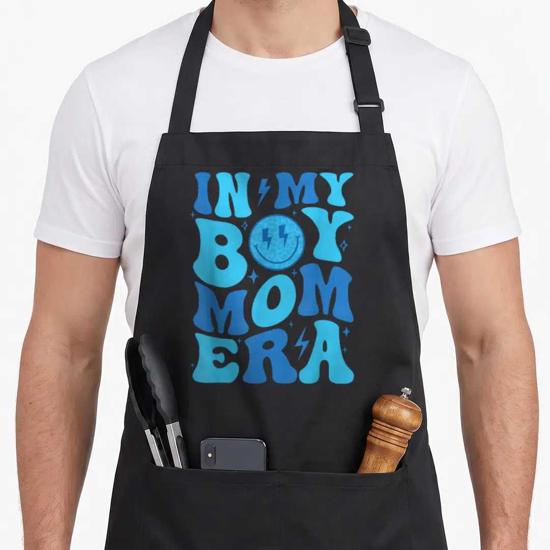 Era mama apron - Blue bubbly text reading IN MY BOY MOM ERA with lightning bolt eyes smiley face and scattered stars — shown on a black cotton canvas apron against a white background