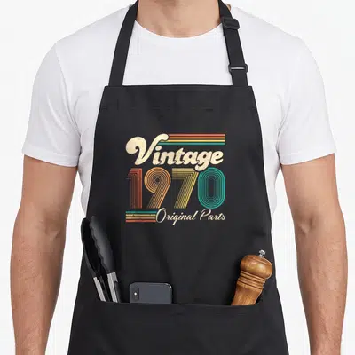 50th bday birthday apron - Retro 1970 typography with cream script and orange yellow teal striped numbers — shown on a black cotton canvas apron against a white background