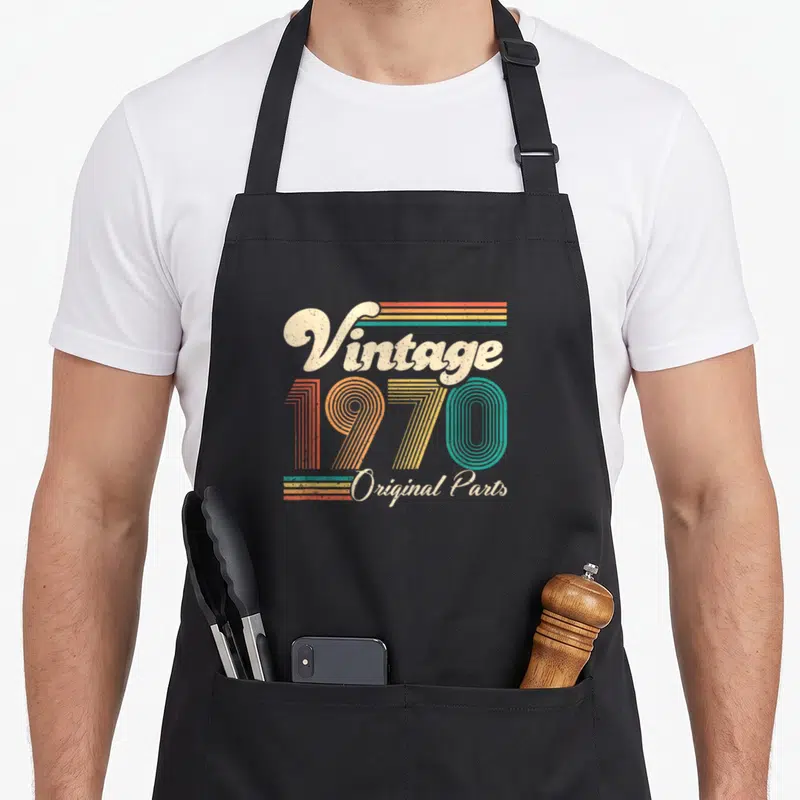 50th bday birthday apron - Retro 1970 typography with cream script and orange yellow teal striped numbers — shown on a black cotton canvas apron against a white background