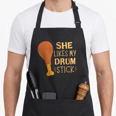 She thanksgiving apron - Cartoon orange chicken drumstick next to stacked text reading SHE LIKES MY DRUM STICK — shown on a black cotton canvas apron against a white background