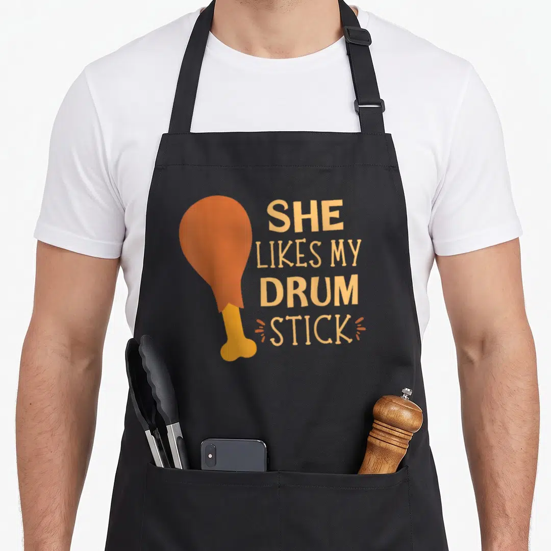 She thanksgiving apron - Cartoon orange chicken drumstick next to stacked text reading SHE LIKES MY DRUM STICK — shown on a black cotton canvas apron against a white background