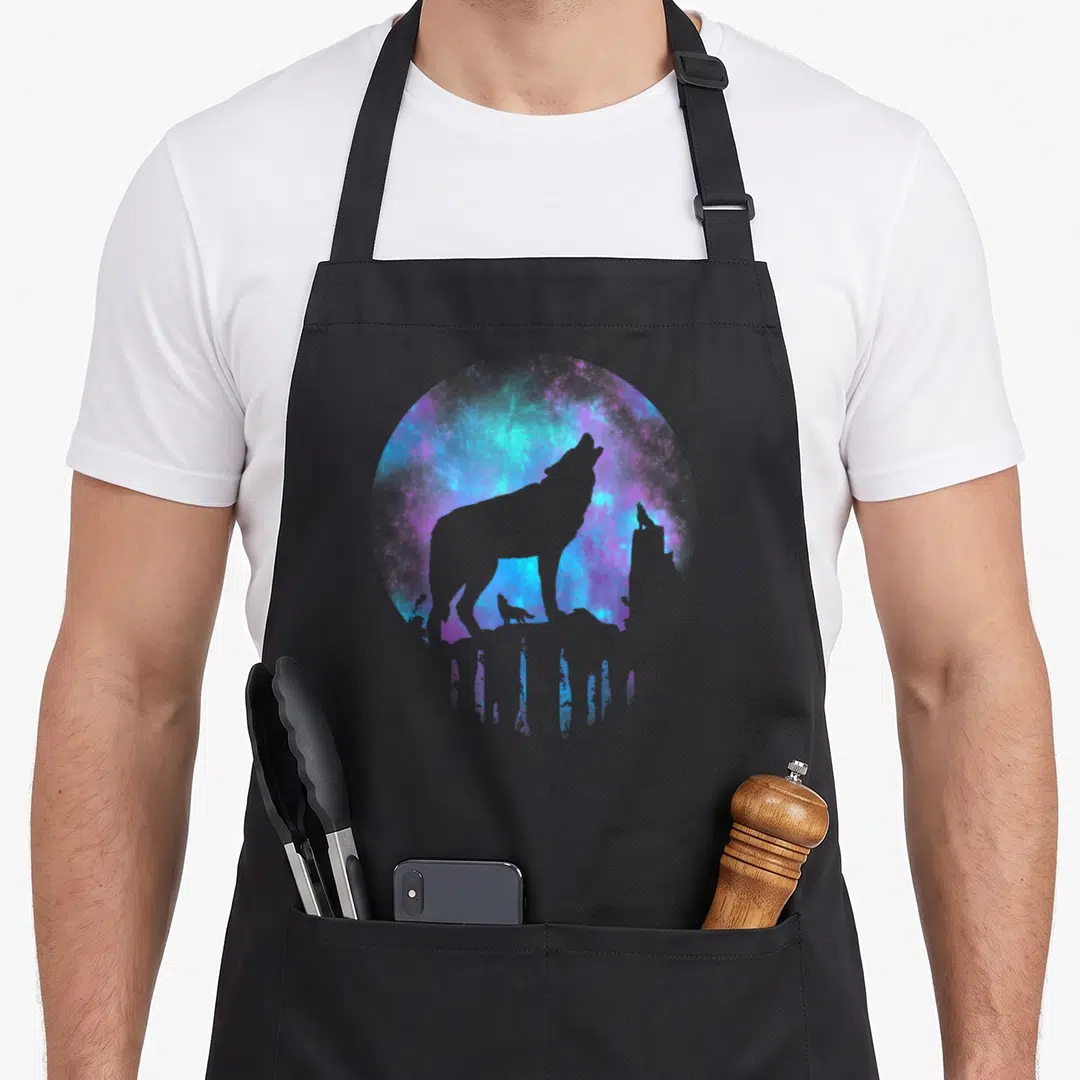 Wolf galaxies apron - Large wolf silhouette and small wolf on rock against blue purple magenta galaxy circle — shown on a black cotton canvas apron against a white background