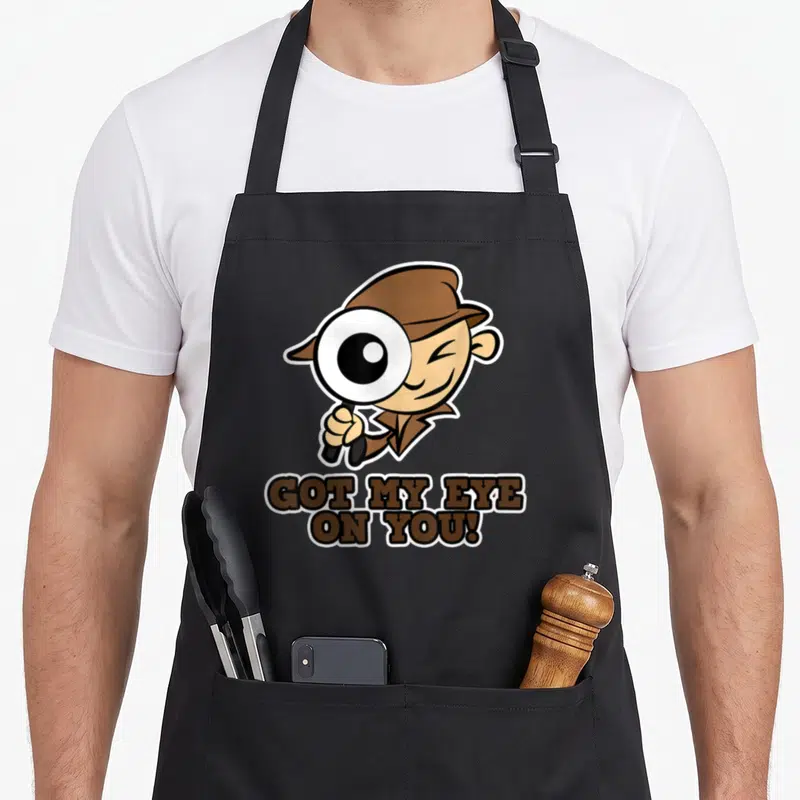 Eye detective apron - Cartoon detective holding magnifying glass above GOT MY EYE ON YOU text — shown on a black cotton canvas apron against a white background