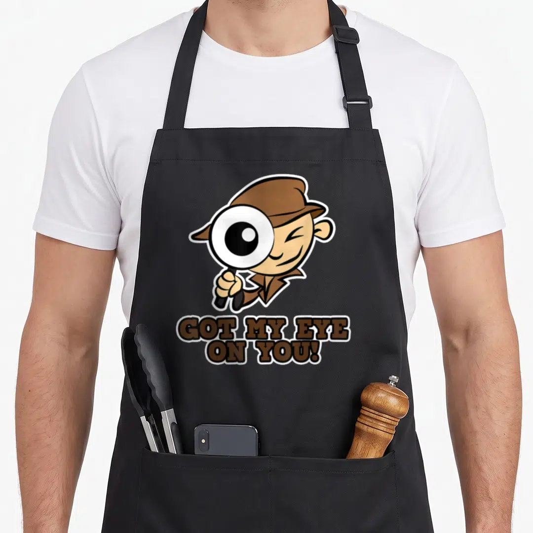 Eye detective apron - Cartoon detective holding magnifying glass above GOT MY EYE ON YOU text — shown on a black cotton canvas apron against a white background