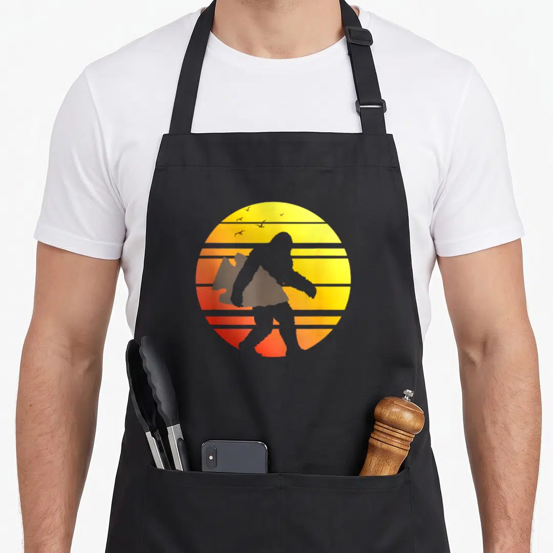 Arrowhead bigfoot apron - Bigfoot silhouette carrying a blocky shape against a striped yellow orange and red retro sun — shown on a black cotton canvas apron against a white background