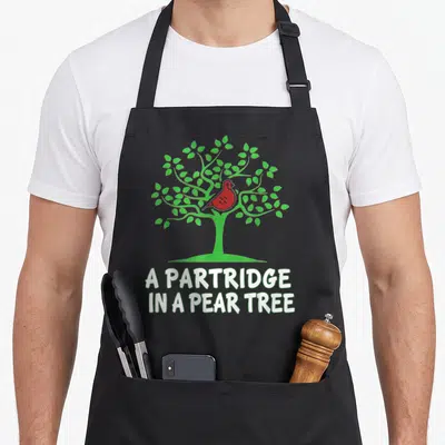 Partridge christmas tree apron - Red cartoon bird in green tree above white text A PARTRIDGE IN A PEAR TREE — shown on a black cotton canvas apron against a white background