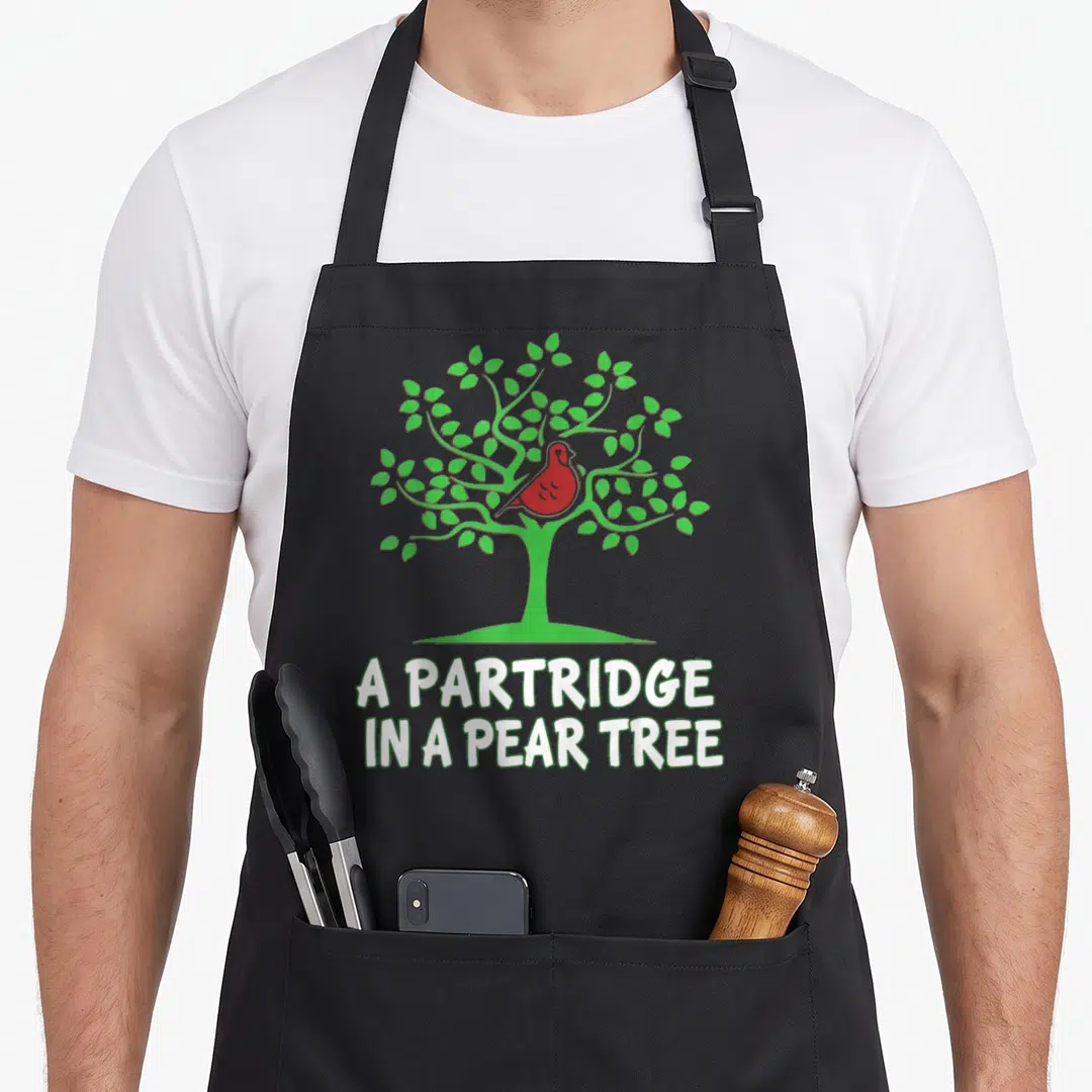 Partridge christmas tree apron - Red cartoon bird in green tree above white text A PARTRIDGE IN A PEAR TREE — shown on a black cotton canvas apron against a white background