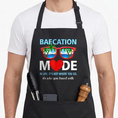 Trip couple apron - Baecation Mode text with red sunglasses showing palm trees and ocean waves — shown on a black cotton canvas apron against a white background