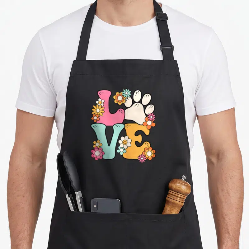 Groovy funny cat aprons - Colorful block letters spelling LOVE with a white paw print and scattered cartoon flowers — shown on a black cotton canvas apron against a white background