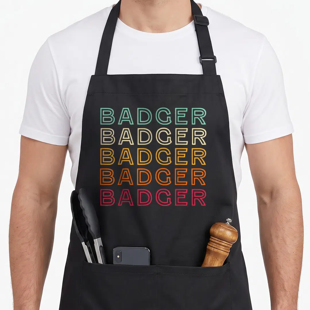 Funny badger apron - Stacked BADGER text in teal beige yellow orange red outlines — shown on a black cotton canvas apron against a white background