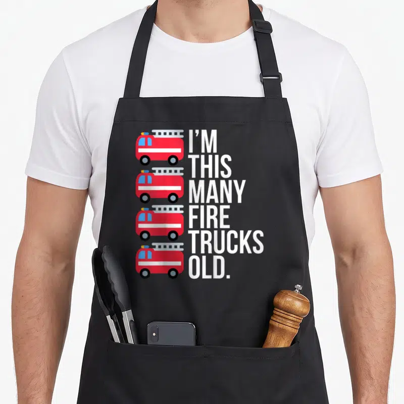 Many fire truck apron - Four red fire trucks stacked vertically beside white text reading I'M THIS MANY FIRE TRUCKS OLD — shown on a black cotton canvas apron against a white background