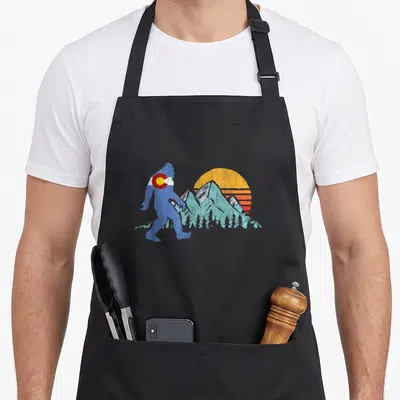 Bigfoot colorado apron - Blue bigfoot silhouette with flag band walking past striped sun and mountains — shown on a black cotton canvas apron against a white background