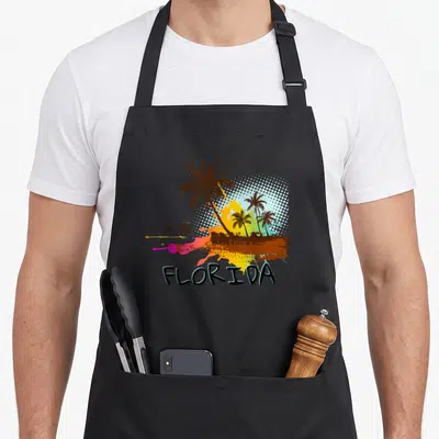 Beach florida apron - Palm trees, dolphins, birds, and FLORIDA text over colorful splatter and halftone dots — shown on a black cotton canvas apron against a white background