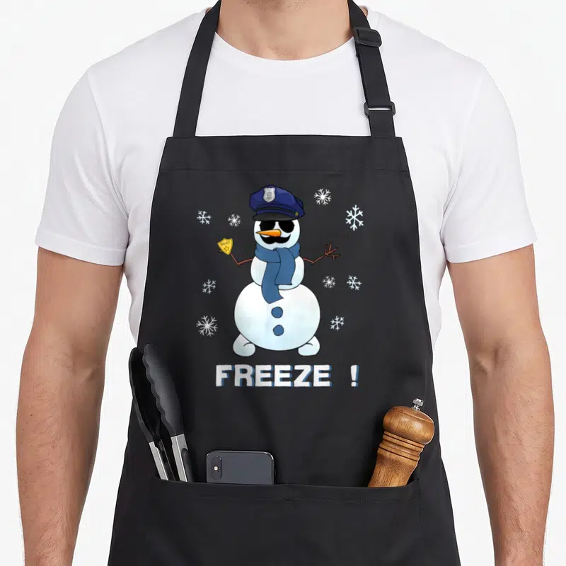 Cop snowman apron - Cartoon snowman in blue police hat holding gold badge with FREEZE ! text and snowflakes — shown on a black cotton canvas apron against a white background