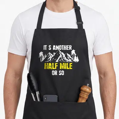 Hiker apron or apron - Distressed white mountains with yellow text reading IT S ANOTHER HALF MILE OR SO — shown on a black cotton canvas apron against a white background