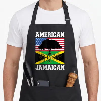 Jamaica american flag apron - Split US and Jamaica flag with black tree and white AMERICAN JAMAICAN text — shown on a black cotton canvas apron against a white background