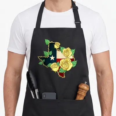 Rose texans apron - Texas map shape filled with blue red flag, outlined by barbed wire with yellow roses and green — shown on a black cotton canvas apron against a white background