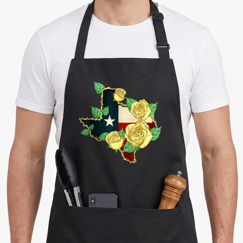 Rose texans apron - Texas map shape filled with blue red flag, outlined by barbed wire with yellow roses and green — shown on a black cotton canvas apron against a white background