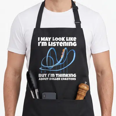 Thinking roller apron - White text reading I may look like I'm listening around a blue cartoon roller coaster track — shown on a black cotton canvas apron against a white background