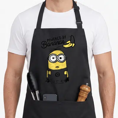 Despicable jook apron - Minion character below arched POWERED BY Bananas text and yellow banana peel — shown on a black cotton canvas apron against a white background